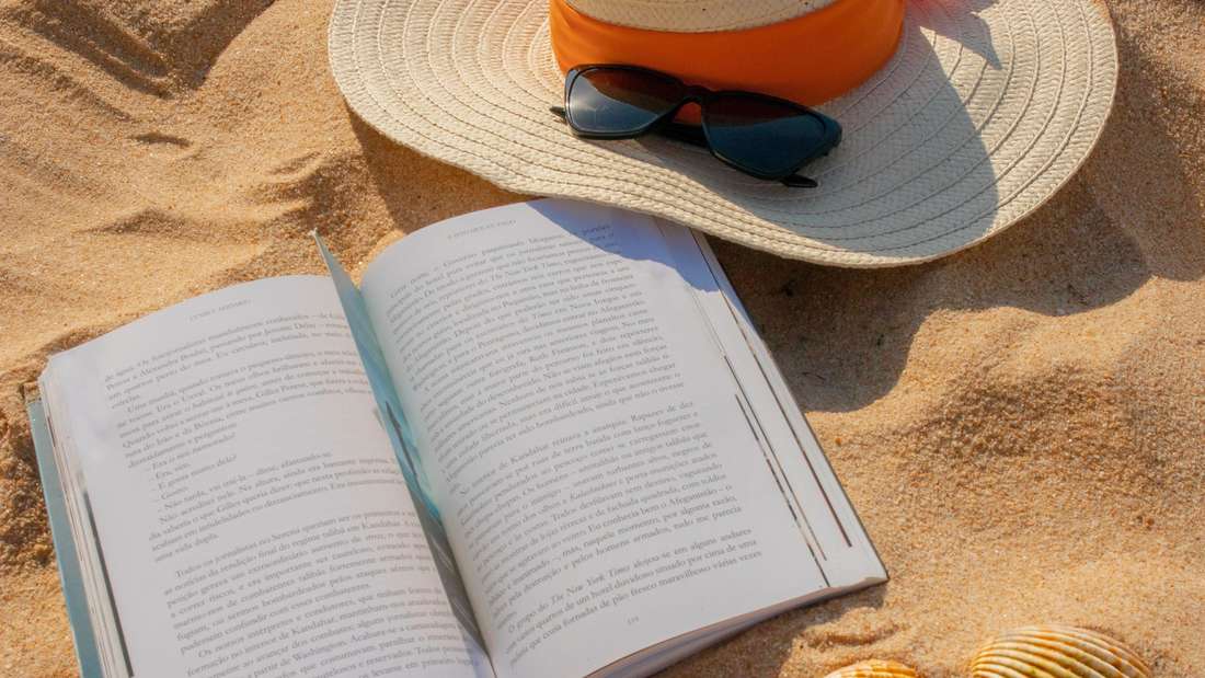 Ein Buch am Strand neben einem Sommerhut (Symbolbild)