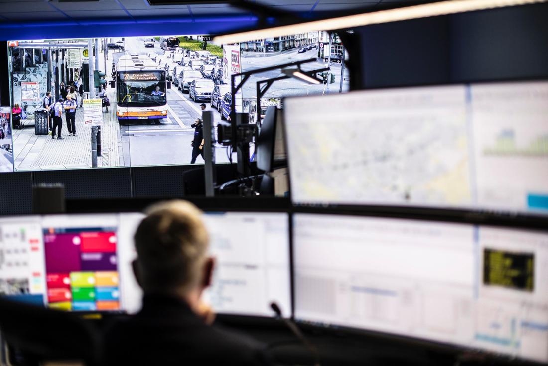 Ein Mitarbeiter der Leitstelle von Eswe Verkehr sitzt an einem Schreibtisch vor mehreren Monitoren. Im Hintergrund laufen Live-Bilder von Haltestellen auf einer Videowand. 