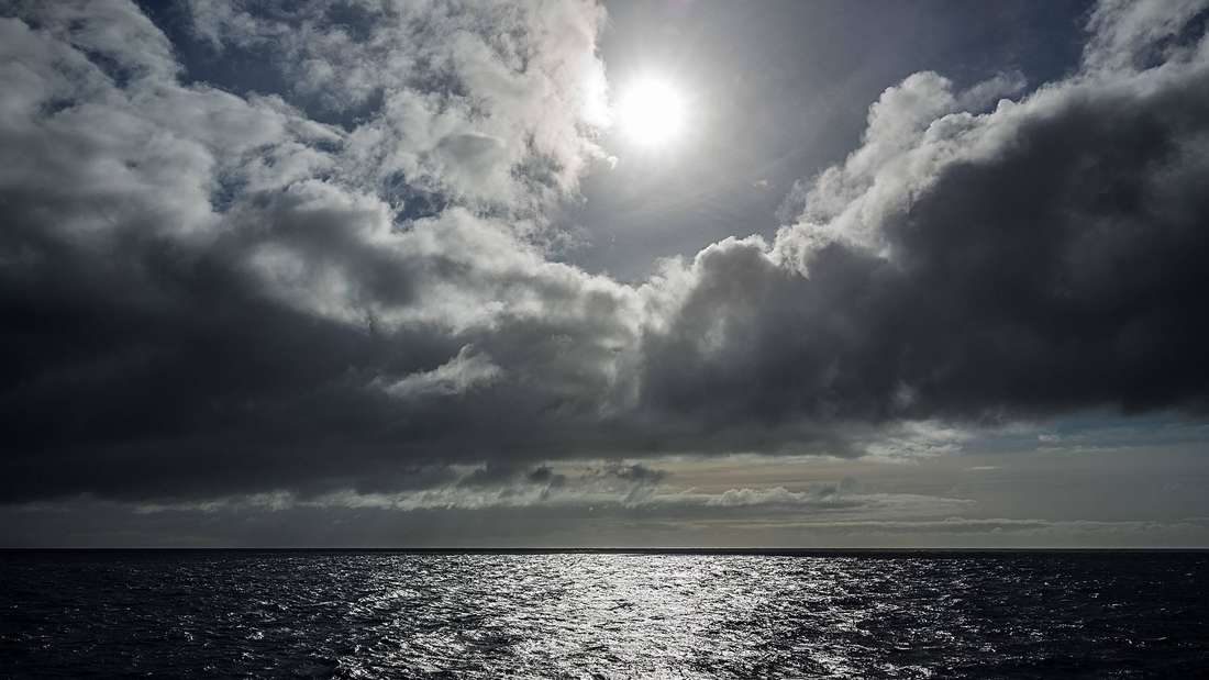 Eine Meeresfläche, darüber Wolken und eine fahle Sonne. 