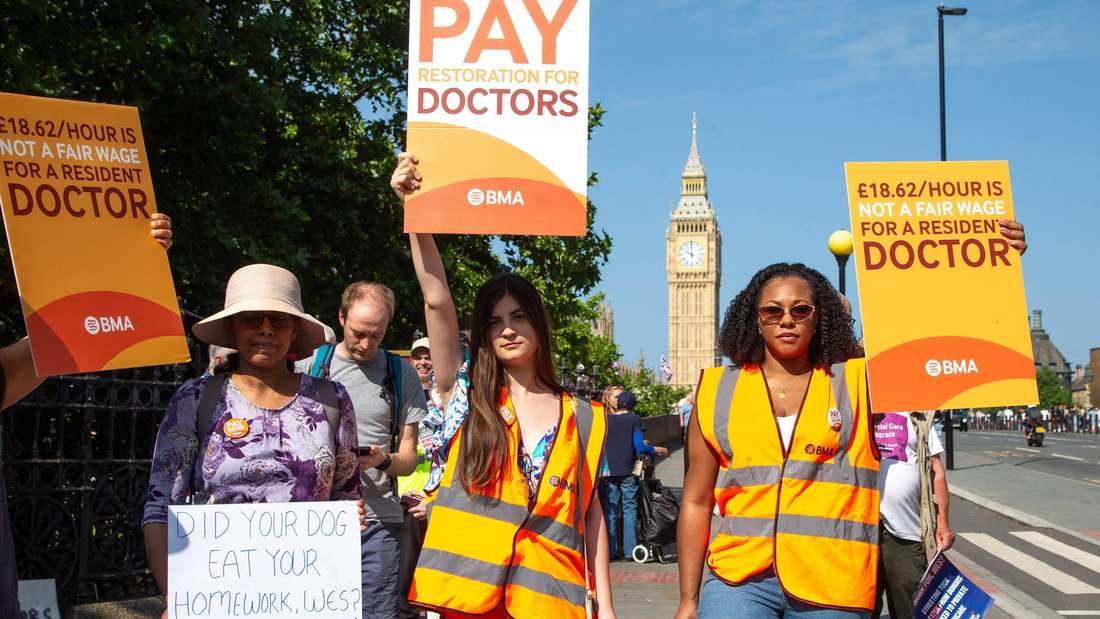 Mehrere Personen mit orangenen Warnwesten, halten Protest-Schilder hoch mit den Aufschriften: Pay restoration for doctors und 18,62 pounds per hour is not a fair wage for a resident doctor.