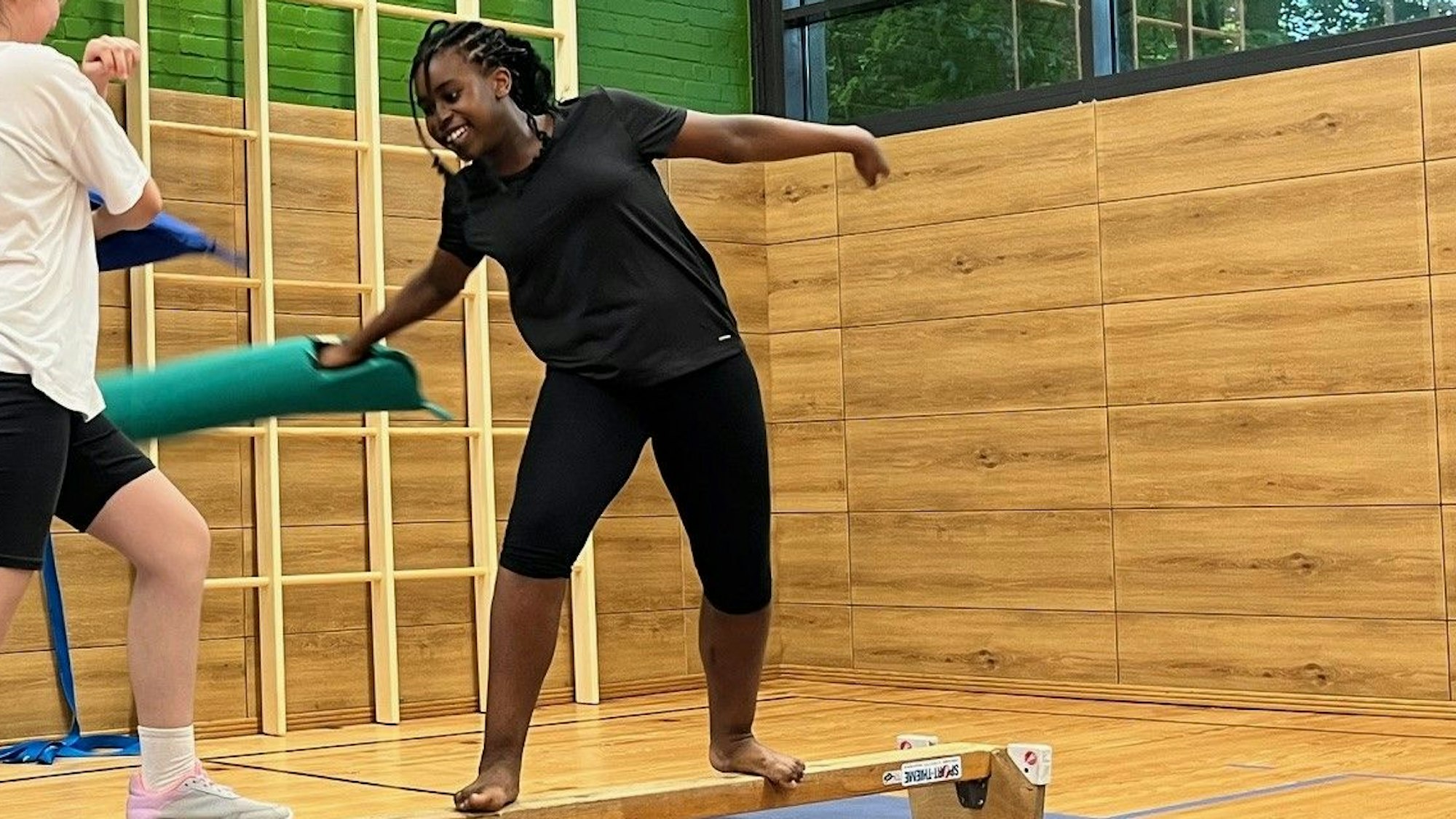 Ein Mädchen steht auf einer Holzbank und versucht, mit einem Bataca, also einem Schläger aus Schaumstoff, ein anderes Mädchen von der Bank zu schubsen.