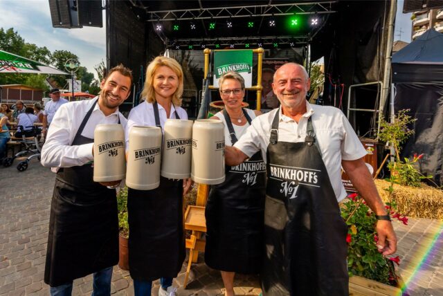 Auf dem Foto sind vier Personen mit Schürzen und Bierkrügen der Marke Brinkhoff’s zu sehen. Sie stehen vor einer Bühne und scheinen an einer Veranstaltung teilzunehmen oder zu arbeiten.