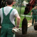 Zwei Mitarbeiter der Kampfmittelbeseitigung stehen neben einem entschärften Bombenblindgänger, der am Haken eines Baufahrzeugs hängt.