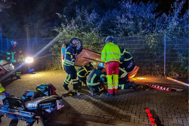 Feuerwehrkräfte stehen und knien vor einem Auto, das auf dem Dach liegt. Sie befreien eine Person.
