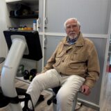 Ein älterer Herr sitzt in Sportkleidung auf einem Ergometer. Im Hintergrund sind Fitnessgeräte und -utensilien zu sehen.
