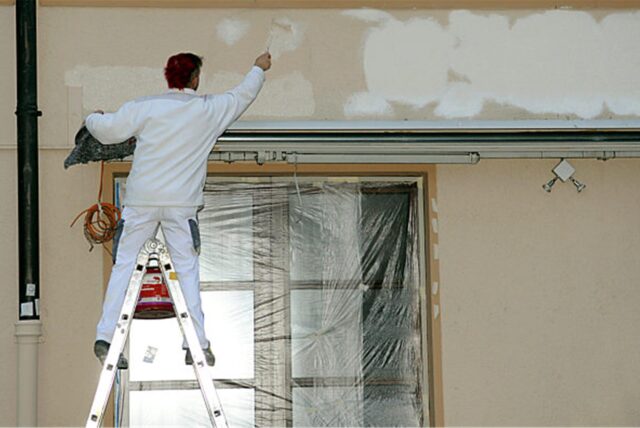 Ein Maler steht auf einer Leiter und renoviert die Außenwand eines Gebäudes. Das Fenster ist mit Folie abgedeckt, während der Maler mit einer Farbrolle weiße Farbe aufträgt.