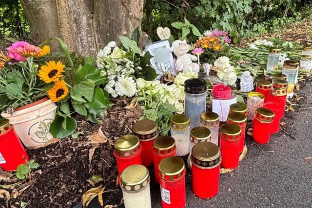 Am Straßenrand sind Blumen, Kerzen und ein Foto als Gedenkstätte an einem Baum platziert. Die Gegenstände säumen den Rand der Fahrbahn, neben einem Gehweg und einer Bushaltestelle.