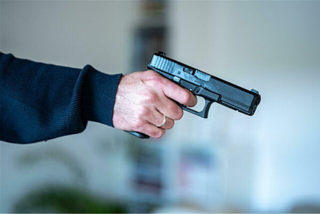 Ein Mann hält eine schwarze Pistole in der Hand. Der Mann trägt einen dunklen Pullover und einen Ring am Finger.