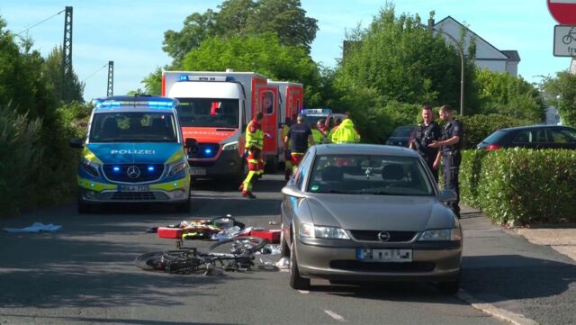 Das Fahrrad des Unfallopfers liegt neben einem Pkw. Im Hintergrund parken mehrere Einsatzfahrzeuge der Feuerwehr und des Rettungsdienstes. Mehrere Rettungskräfte und Polizeibeamte stehen im Hintergrund.