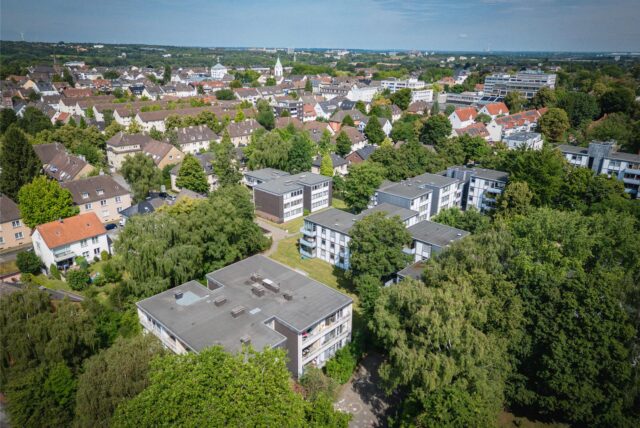 Eine Luftaufnahme zeigt eine Wohngegend mit Mehrfamilienhäusern und Einfamilienhäusern in Dortmund-Hombruch. Die Gebäude sind von viel Grün umgeben, und im Hintergrund ist eine Kirchturmspitze zu sehen.