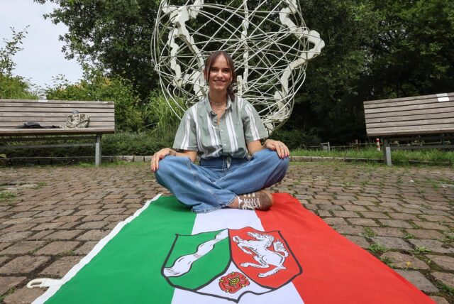 Annika Faust sitzt auf einer NRW-Fahne vor dem Kunstwerk.