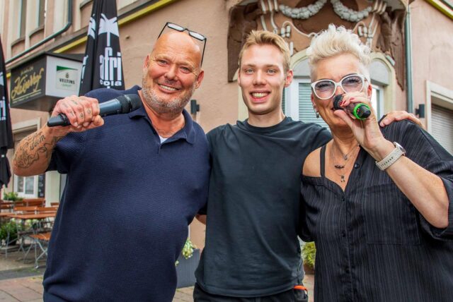Elke Moschny und Dirk Lasarz alias Duo Just4Fun stehen mit einem Mikrofon in der Hand in der Innenstadt von Herten. Tontechniker Robin Pospiech steht zwischen ihnen.