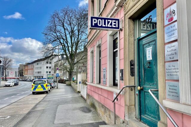 Eine Außenaufnahme eines Polizeireviers an einem sonnigen Tag. Im Vordergrund ist der Eingang des Gebäudes zu sehen, während im Hintergrund ein Streifenwagen geparkt ist.