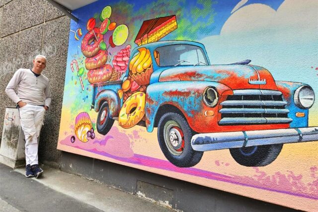 Ein Mann posiert vor einem Wandgemälde, das einen Oldtimer zeigt, der mit Süßigkeiten beladen ist. Das Kunstwerk ist farbenfroh und detailreich gestaltet.