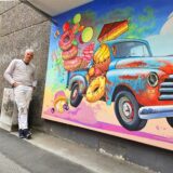 Ein Mann posiert vor einem Wandgemälde, das einen Oldtimer zeigt, der mit Süßigkeiten beladen ist. Das Kunstwerk ist farbenfroh und detailreich gestaltet.