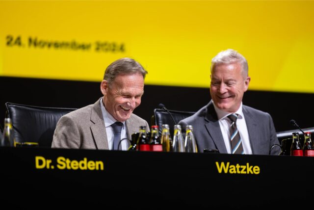 Hans-Joachim Watzke und Thomas Treß sitzen auf einem Podium und lachen.