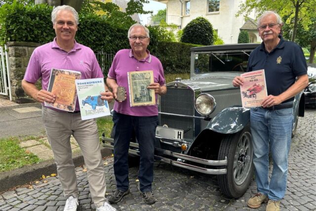 Drei Herren präsentieren historische Dokumente und Memorabilien vor einem Oldtimer. Die Gegenstände beziehen sich auf Automobilveranstaltungen und die Automobilgeschichte.