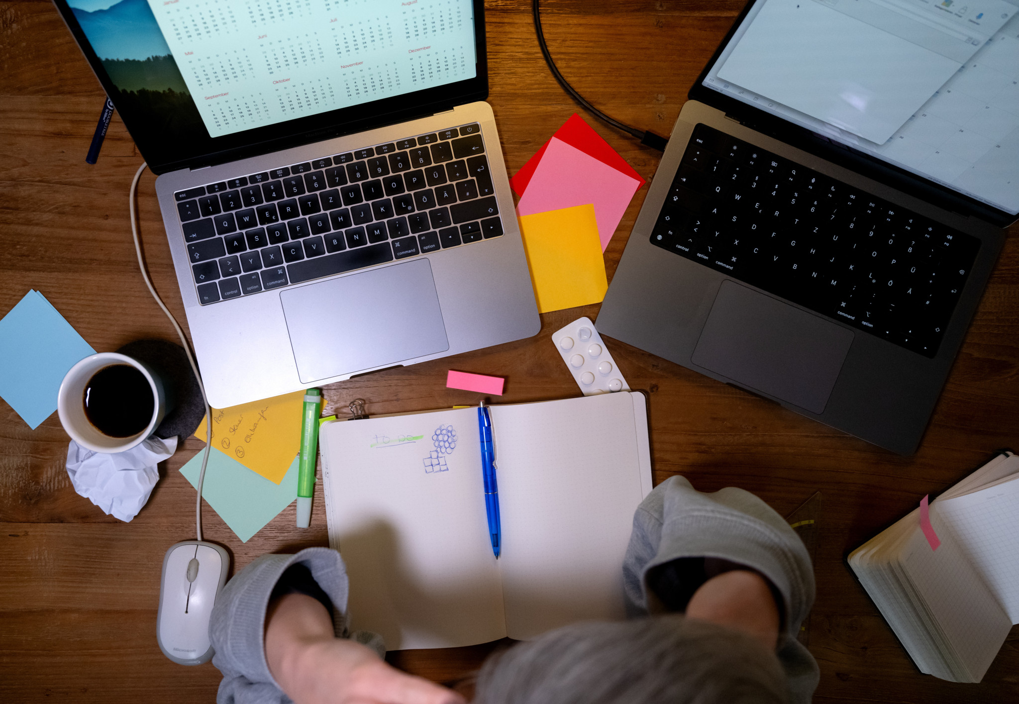Eine Person mit grauen Haaren hält den Kopf in den Händen, während sie an einem Schreibtisch mit zwei Laptops, Notizblöcken und Post-its sitzt. Thema ADHS bei Erwachsenen.