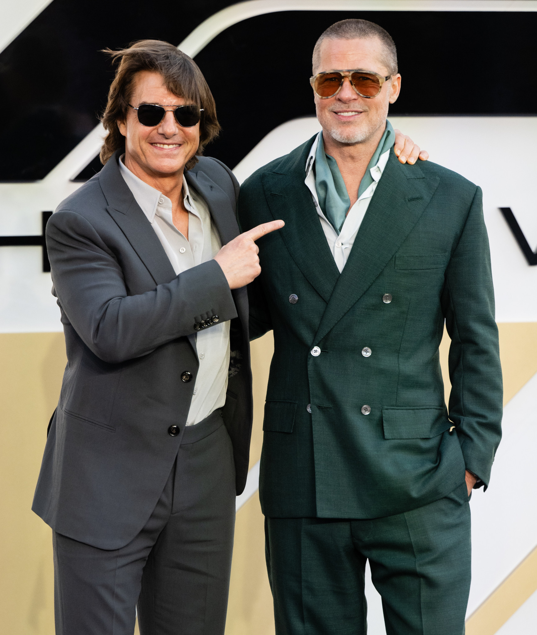 Tom Cruise zeigt auf Brad Pitt bei der Premiere von ’F1: The Movie’ in London, beide in eleganten Anzügen und Sonnenbrillen.