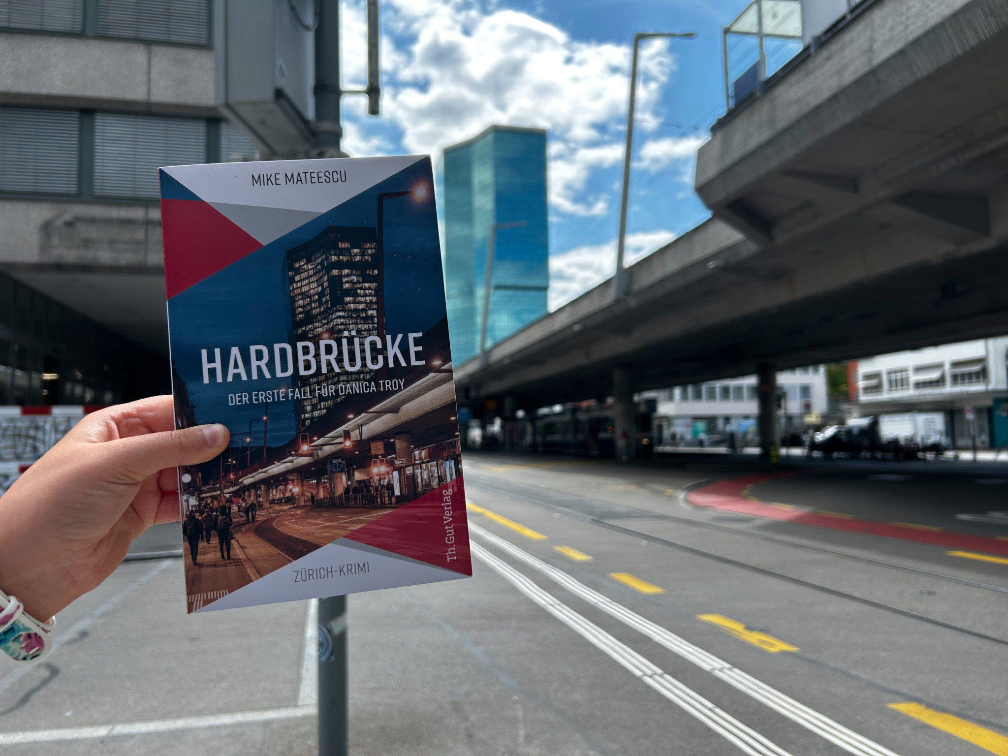 Eine Person hält ein Buch mit dem Titel ’Hardbrücke: Der erste Fall für Corinna Troy’ vor einem modernen Gebäude in Zürich.
