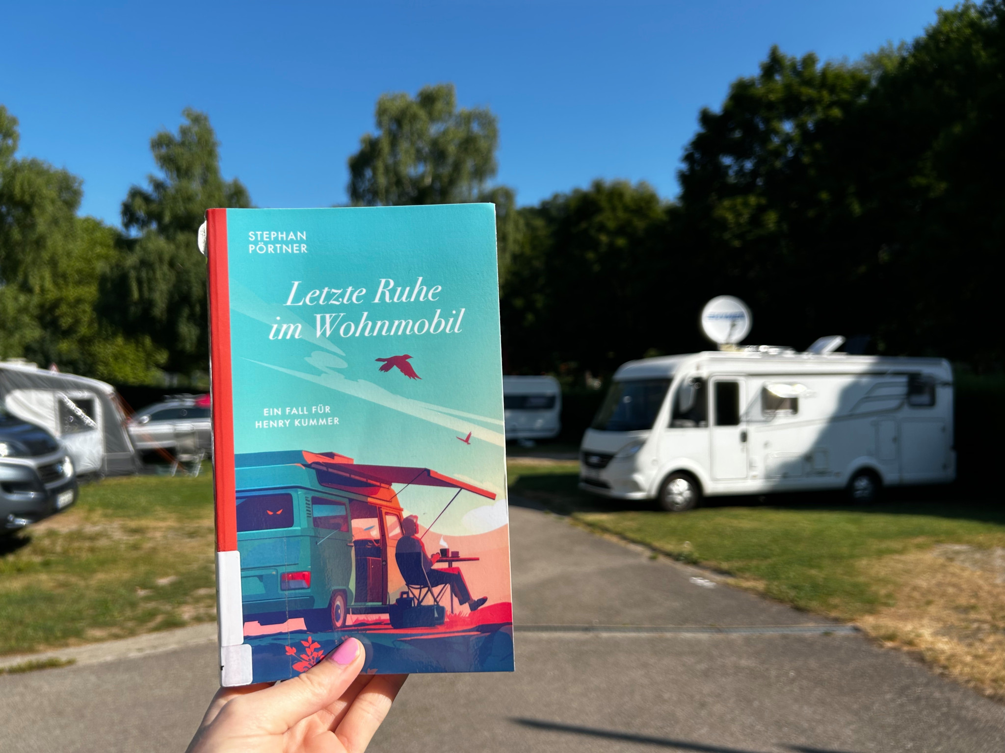 Buch mit dem Titel ’Letzte Ruhe im Wohnmobil’ von Stephan Pörtner vor geparkten Wohnmobilen im Freien gehalten.