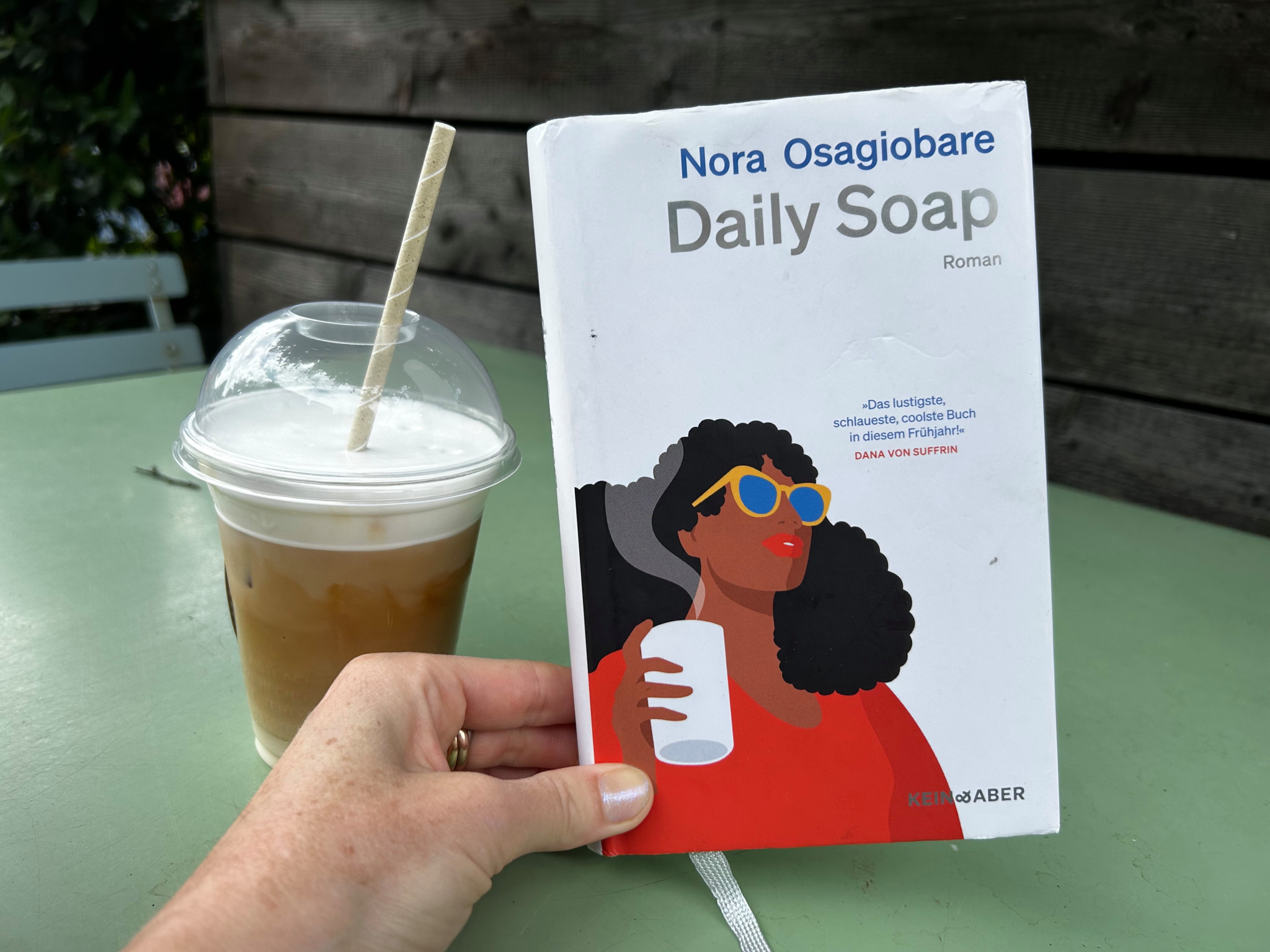 Buch ’Daily Soap’ von Nora Osagiobare neben einem Eiskaffee auf einem Tisch.