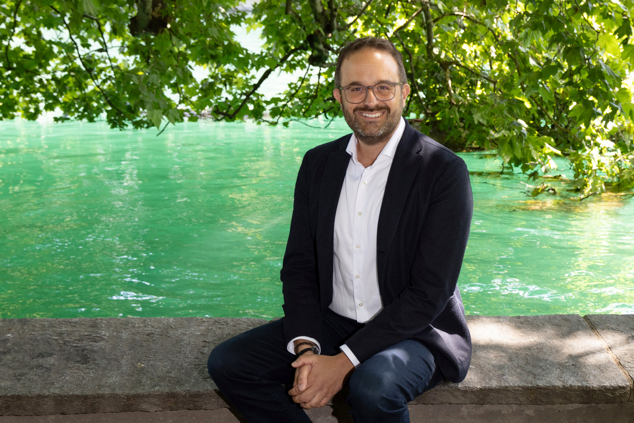 Lukas Geiger sitzt lächelnd vor grünlichem Wasser in Zürich, umgeben von Bäumen, und spricht über sein Leben mit ADHS.