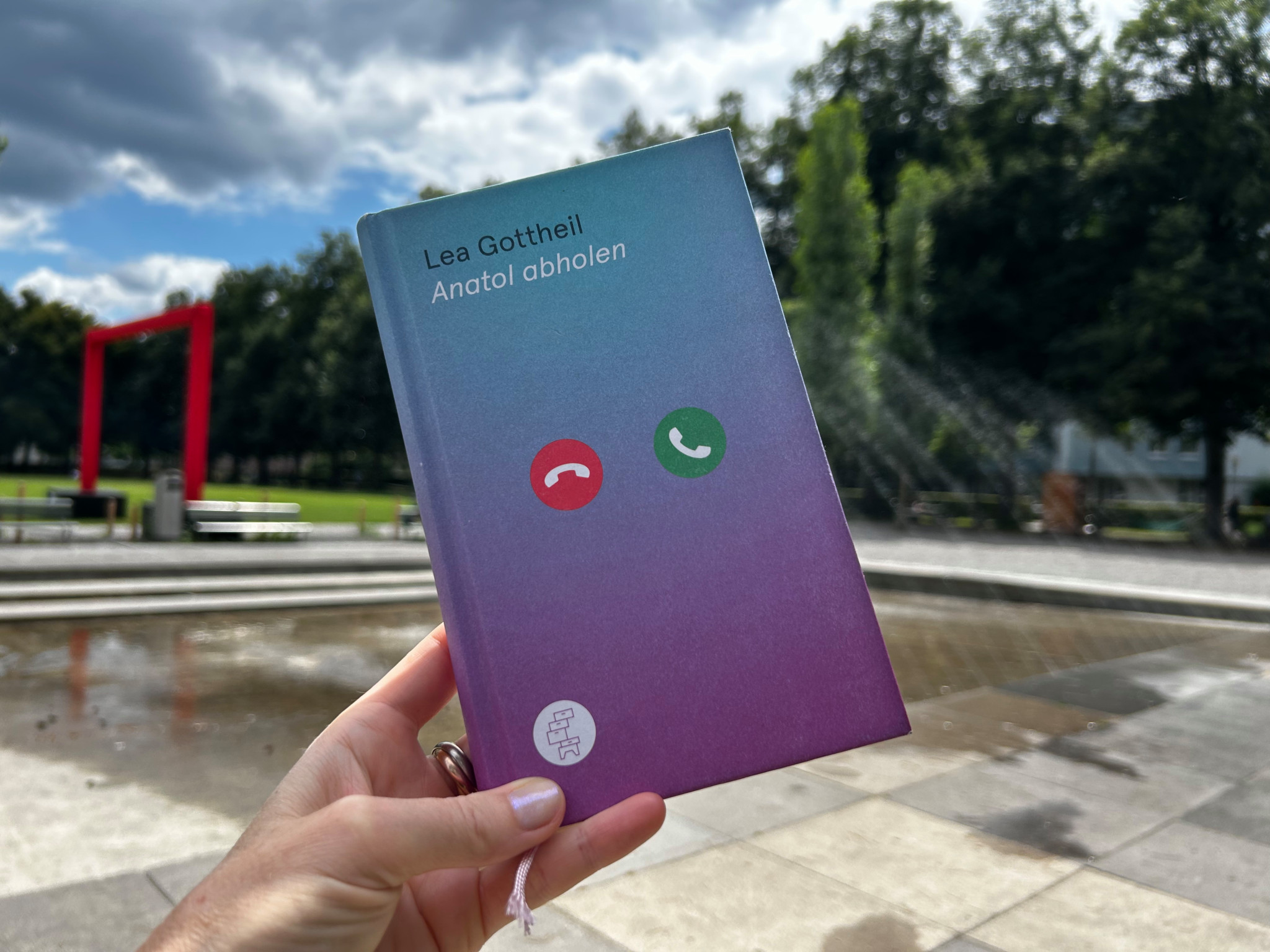 Nahaufnahme eines Buches mit dem Titel ’Anatol abholen’ von Lea Gottheil vor einem Brunnen im Freien gehalten.