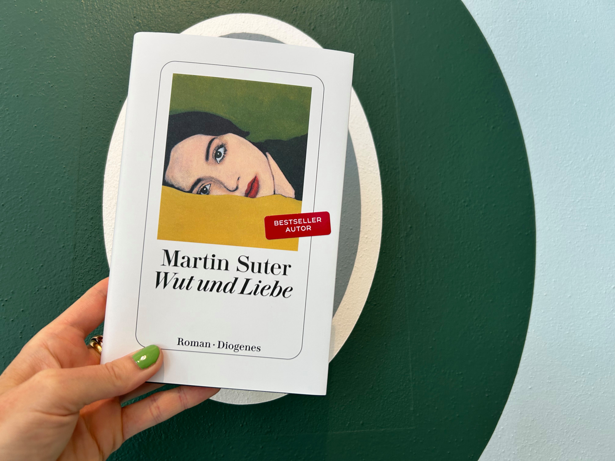 Eine Hand hält das Buch ’Wut und Liebe’ von Martin Suter mit einem Aufkleber ’Bestseller Autor’ auf dem Cover, vor einem grünen und blauen Hintergrund.
