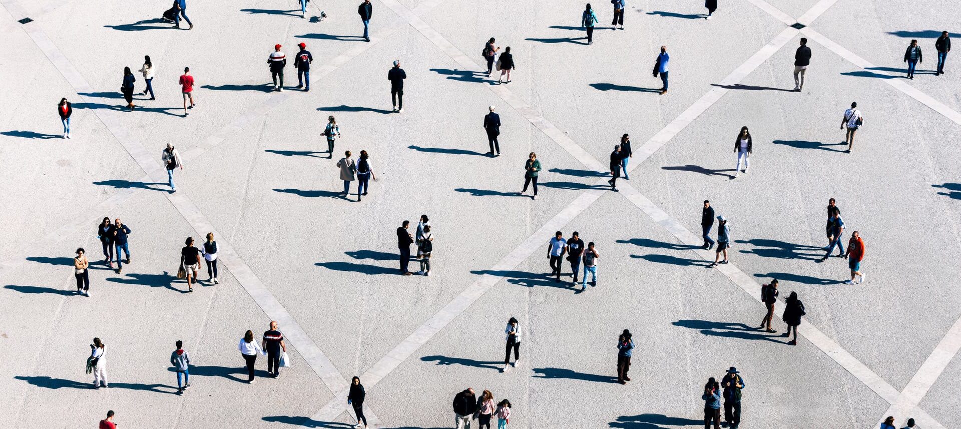 Eine große, offene Fläche mit vielen Menschen, die in kleinen Gruppen oder allein stehen und gehen. Die Szene ist aus der Vogelperspektive aufgenommen, wodurch geometrische Muster auf dem Boden sichtbar werden. Die Personen werfen lange Schatten. Die Kleidung der Menschen ist vielfältig, was auf eine alltägliche, urbane Umgebung schließen lässt.