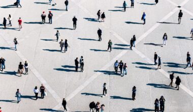 Eine große, offene Fläche mit vielen Menschen, die in kleinen Gruppen oder allein stehen und gehen. Die Szene ist aus der Vogelperspektive aufgenommen, wodurch geometrische Muster auf dem Boden sichtbar werden. Die Personen werfen lange Schatten. Die Kleidung der Menschen ist vielfältig, was auf eine alltägliche, urbane Umgebung schließen lässt.