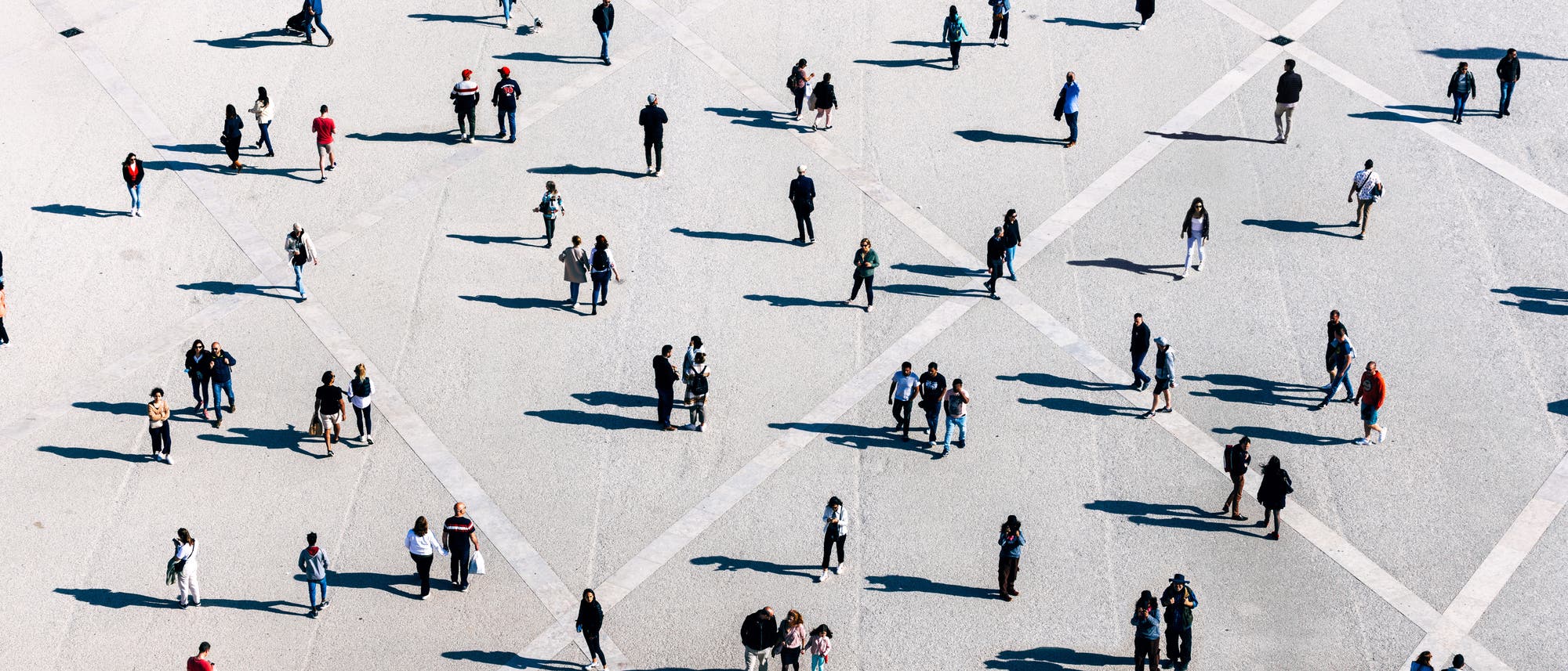 Eine große, offene Fläche mit vielen Menschen, die in kleinen Gruppen oder allein stehen und gehen. Die Szene ist aus der Vogelperspektive aufgenommen, wodurch geometrische Muster auf dem Boden sichtbar werden. Die Personen werfen lange Schatten. Die Kleidung der Menschen ist vielfältig, was auf eine alltägliche, urbane Umgebung schließen lässt.