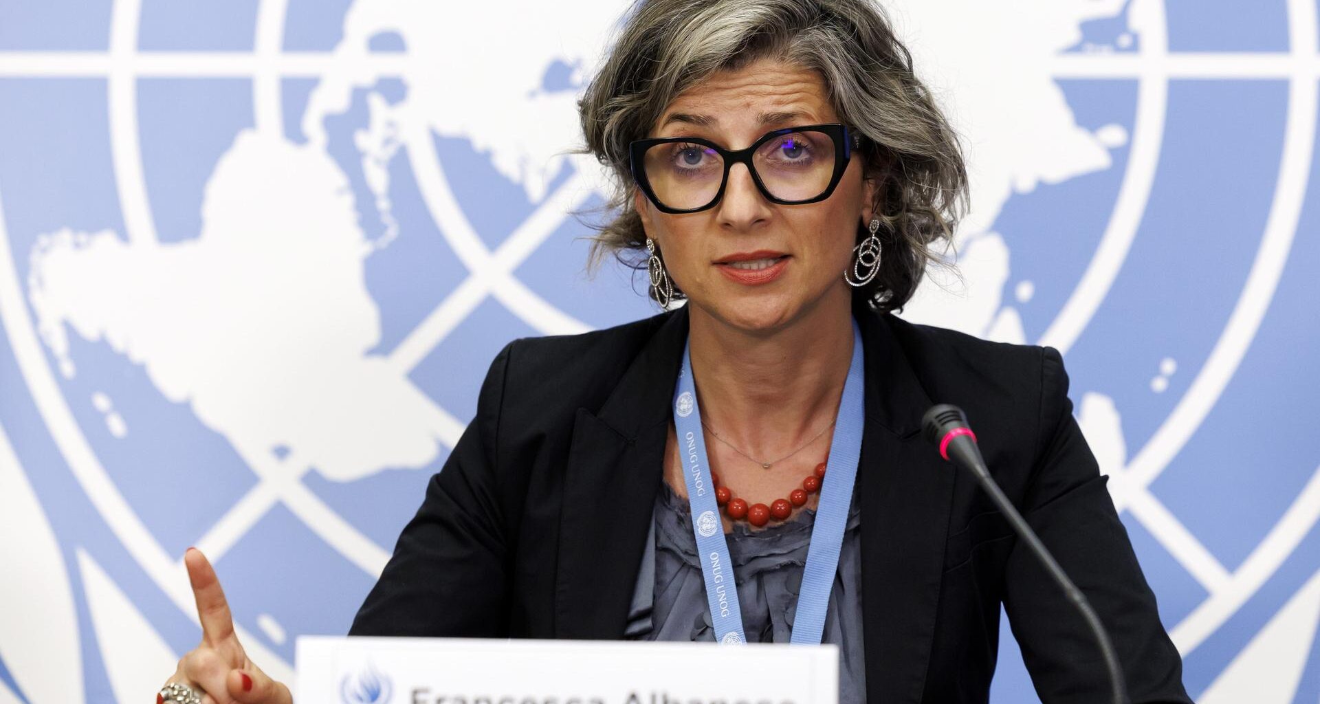 Francesca Albanese sitzt an einem Tisch, spricht in ein Mikrofon und hebt dabei den Zeigefinger der rechten Hand. Im Hintergrund ist eine Weltkarte zu sehen.