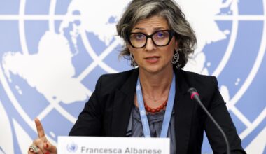 Francesca Albanese sitzt an einem Tisch, spricht in ein Mikrofon und hebt dabei den Zeigefinger der rechten Hand. Im Hintergrund ist eine Weltkarte zu sehen.