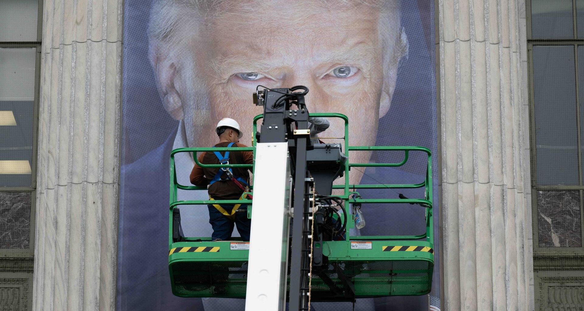 Donald Trump als Plakat. Seine Augen sind hinter einer Plattform zu erkennen, auf der ein Arbeiter steht. Vermutlich bringt dieser gerade das überlebensgroße Banner an