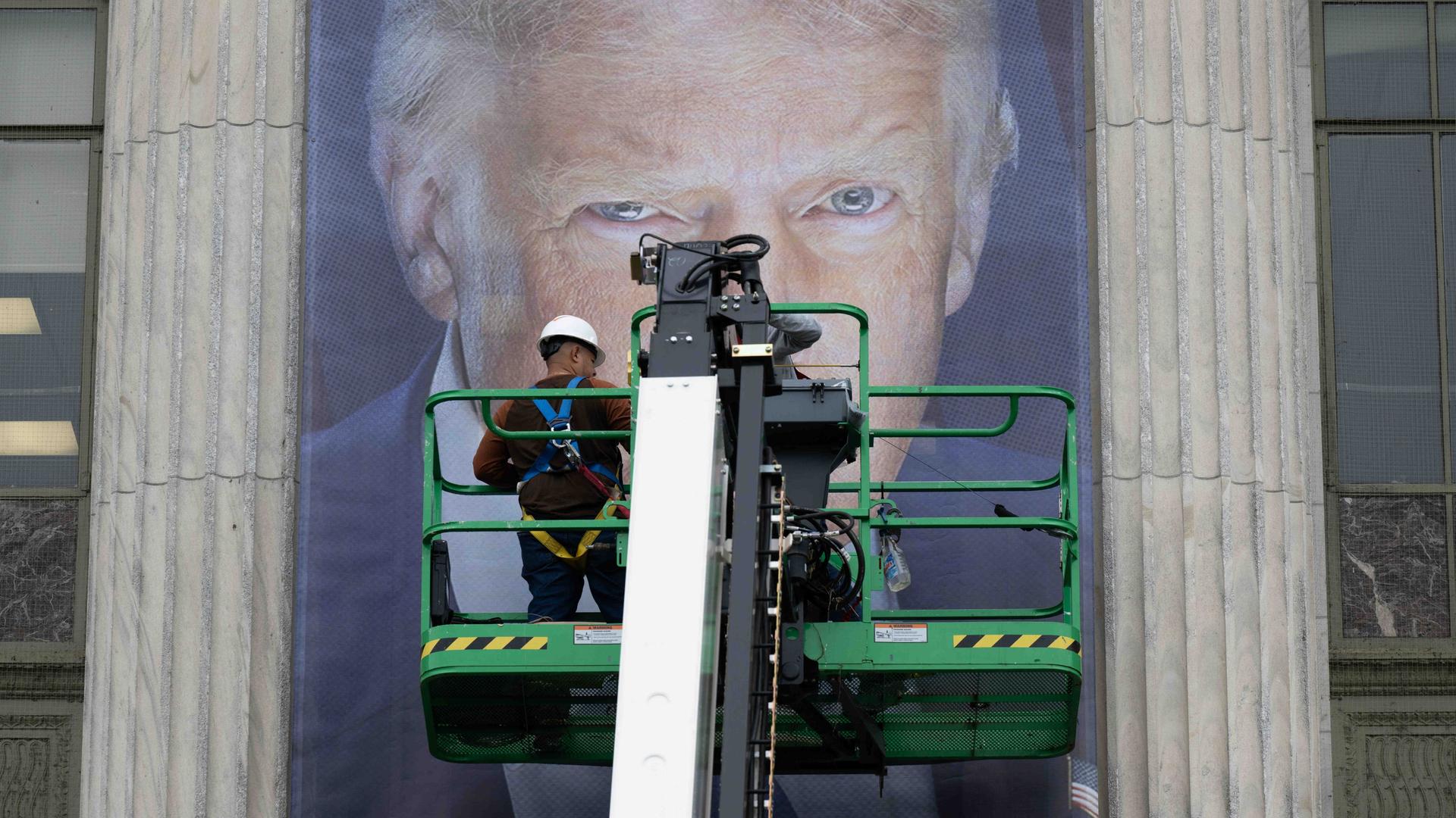 Donald Trump als Plakat. Seine Augen sind hinter einer Plattform zu erkennen, auf der ein Arbeiter steht. Vermutlich bringt dieser gerade das überlebensgroße Banner an