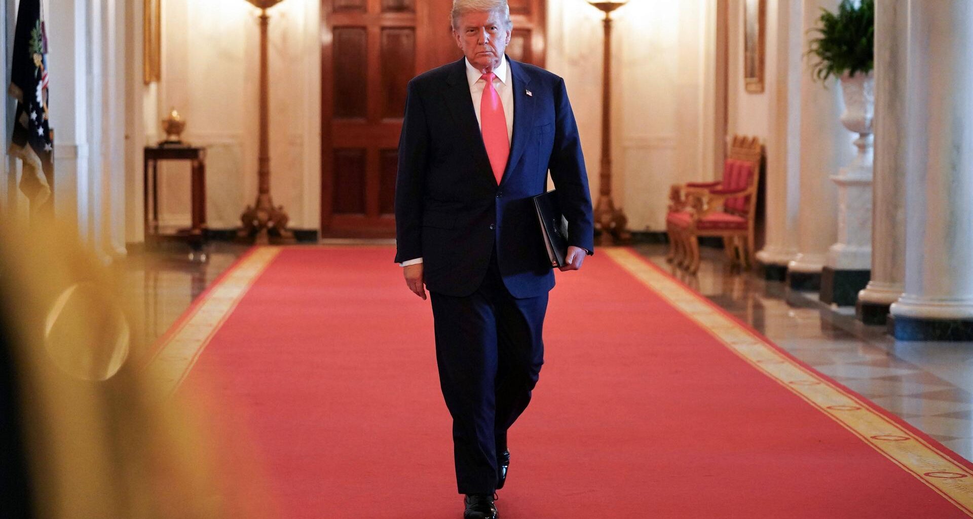 Donald Trump mit Aktenmappe unter dem Arm auf dem roten teppich in einem Korridor des Weißen Hauses