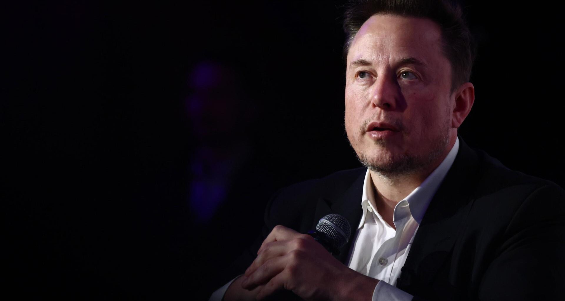 Elon Musk trägt einen Anzugu und sitzt vor einem dunklen Hintergrund, in den hält er ein Mikrofon.