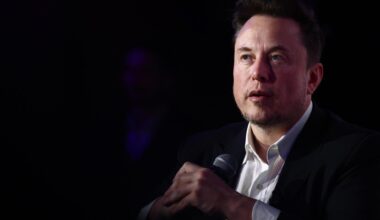 Elon Musk trägt einen Anzugu und sitzt vor einem dunklen Hintergrund, in den hält er ein Mikrofon.