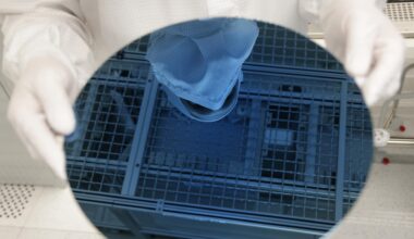 Ein Mitarbeiter in weißer Schutzkleidung hält einen Wafer in den Händen, eine runde, bläuliche Glasscheibe.