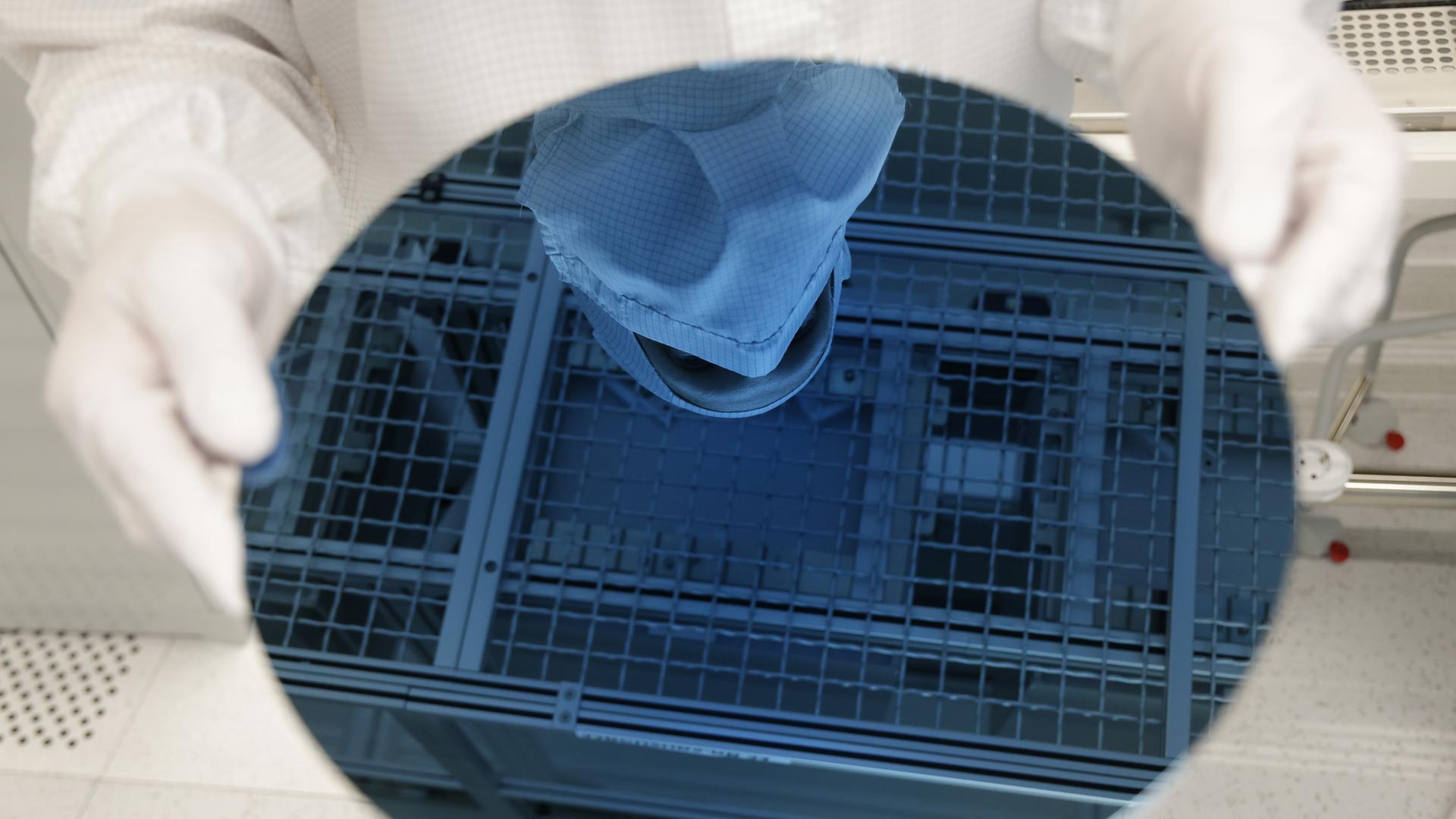 Ein Mitarbeiter in weißer Schutzkleidung hält einen Wafer in den Händen, eine runde, bläuliche Glasscheibe.