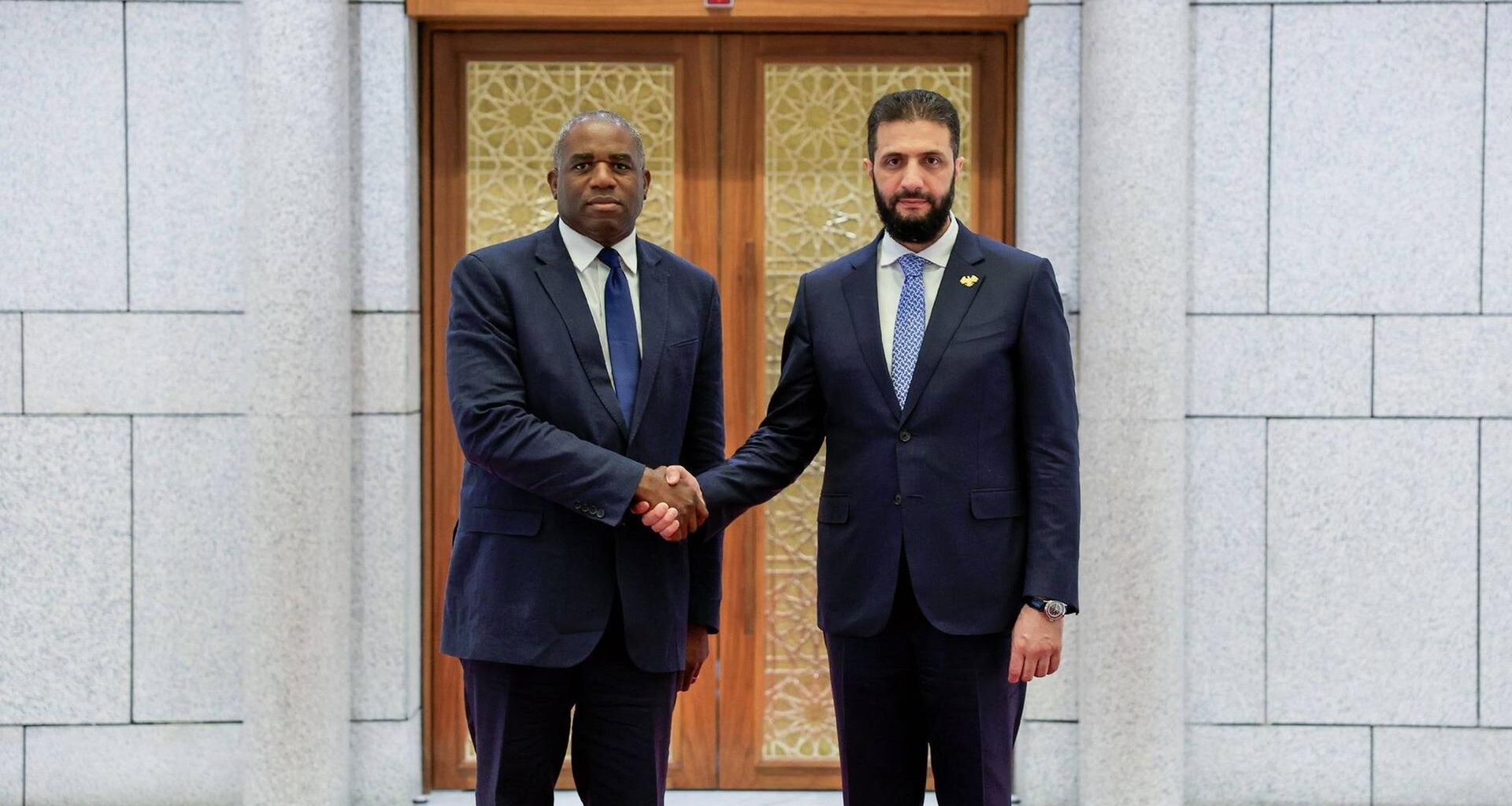 Ahmed al-Sharaa und David Lammy schütteln sich die Hand und schauen in die Kamera.
