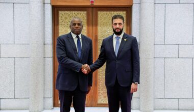 Ahmed al-Sharaa und David Lammy schütteln sich die Hand und schauen in die Kamera.