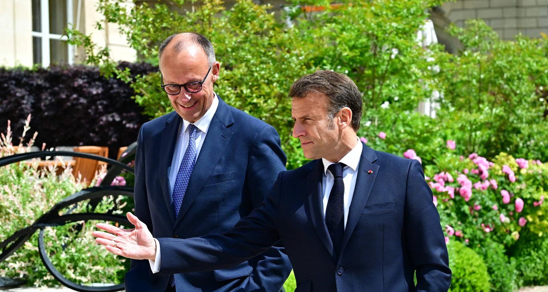 Friedrich Merz (links) und Emmanuel Macron (rechts) laufen gutgelaunt in dunklen Anzugen durch einen Park