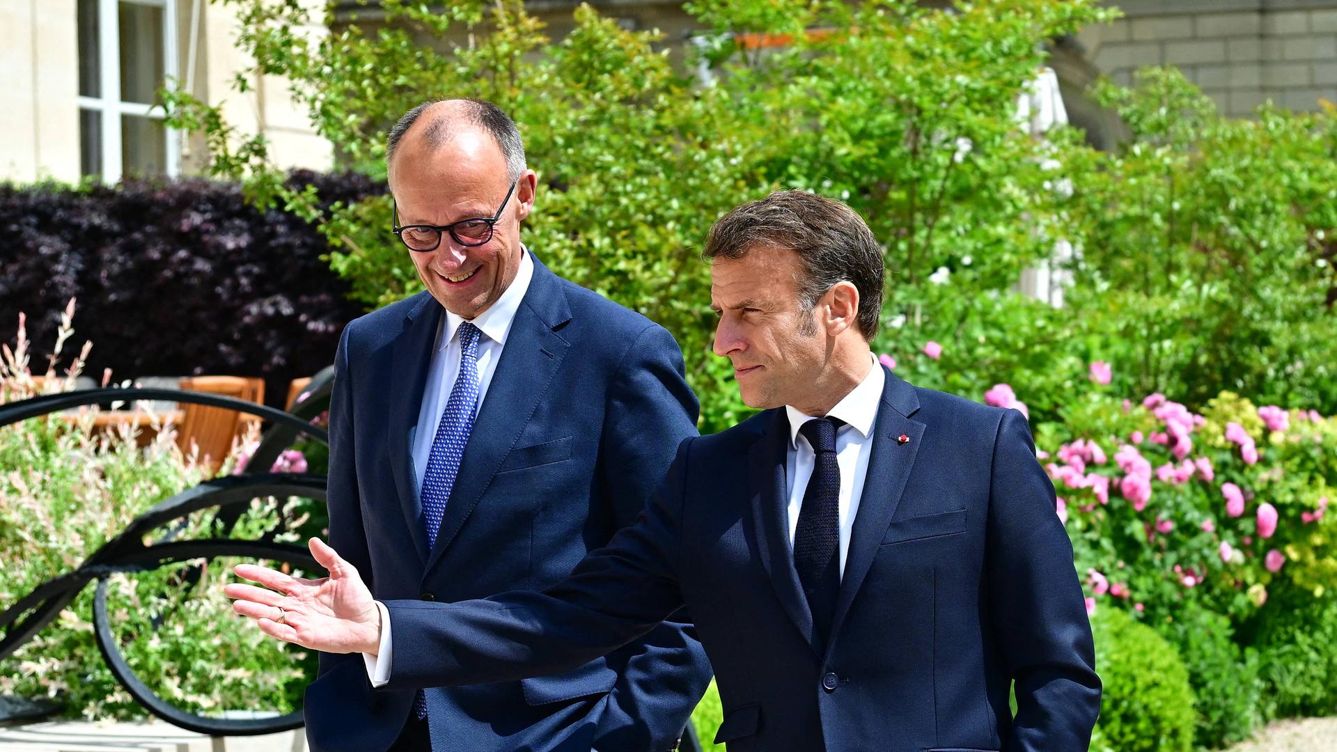 Friedrich Merz (links) und Emmanuel Macron (rechts) laufen gutgelaunt in dunklen Anzugen durch einen Park.