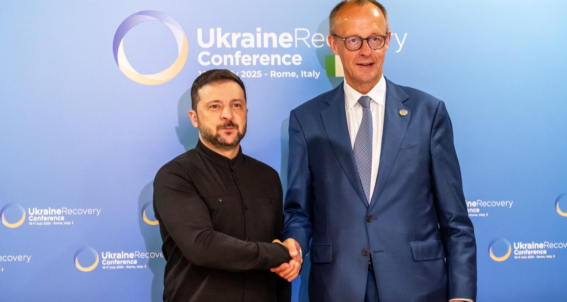 Friedrich Merz und Wolodymyr Selenskyj geben sich die Hand. Im Hintergrund ist auf einer Stellwand das Logo der Wiederaufbaukonferenz für die Ukraine zu sehen.