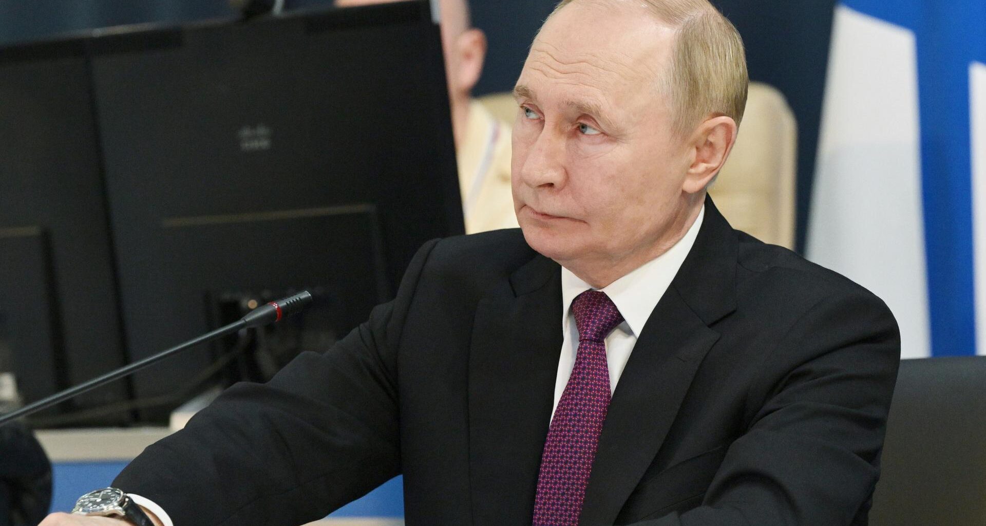 Putin sitzt an einem Mikrofon, im Hintergrund mehrere Bildschirme