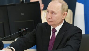 Putin sitzt an einem Mikrofon, im Hintergrund mehrere Bildschirme