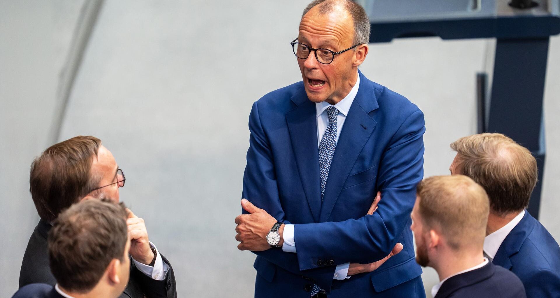 Bundeskanzler Merz spricht im Bundestag mit Ministern von der SPD und der CDU. Sie stehen im Bundestag. Merz hat die Arme verschränkt und den Mund weit aufgerissen.