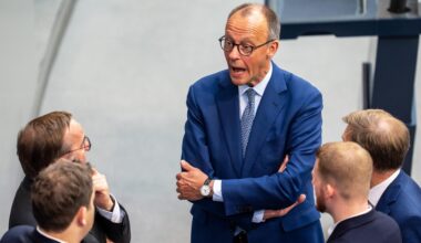 Bundeskanzler Merz spricht im Bundestag mit Ministern von der SPD und der CDU. Sie stehen im Bundestag. Merz hat die Arme verschränkt und den Mund weit aufgerissen.